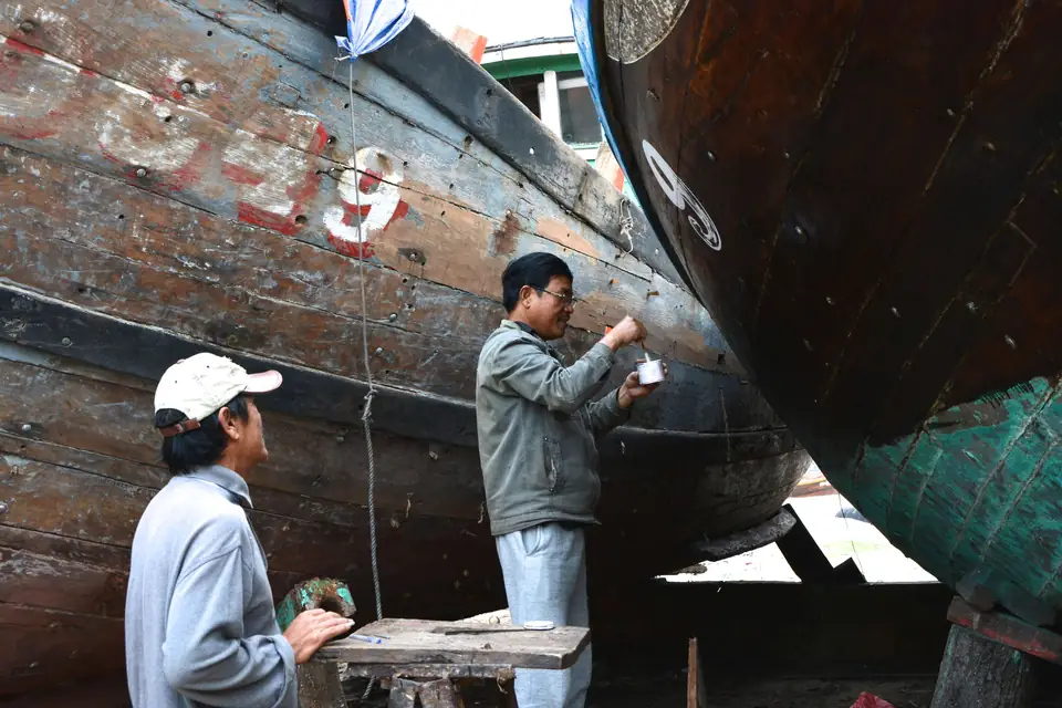 Two people fairing the hull of a large wooden boat in the yard